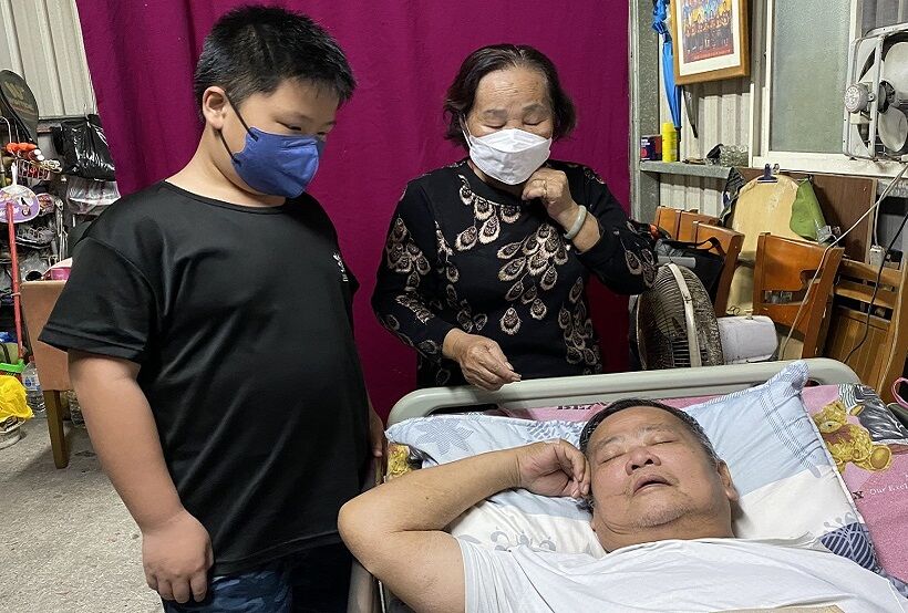 59歲的單親爸阮勝富（阿富）罹肝癌又因染疫咳到傷口裂開再度住院，出院後仍虛弱躺在床上緊閉雙眼，年近80歲老母阿英（圖後方）十分擔心阿富身體，更操心阿富病後全家只靠阿英姨4千多元國民年金不知如何過日子，而且阿富兒子阿駿（圖左），目前才11歲就讀小學五年級。王雪玲攝