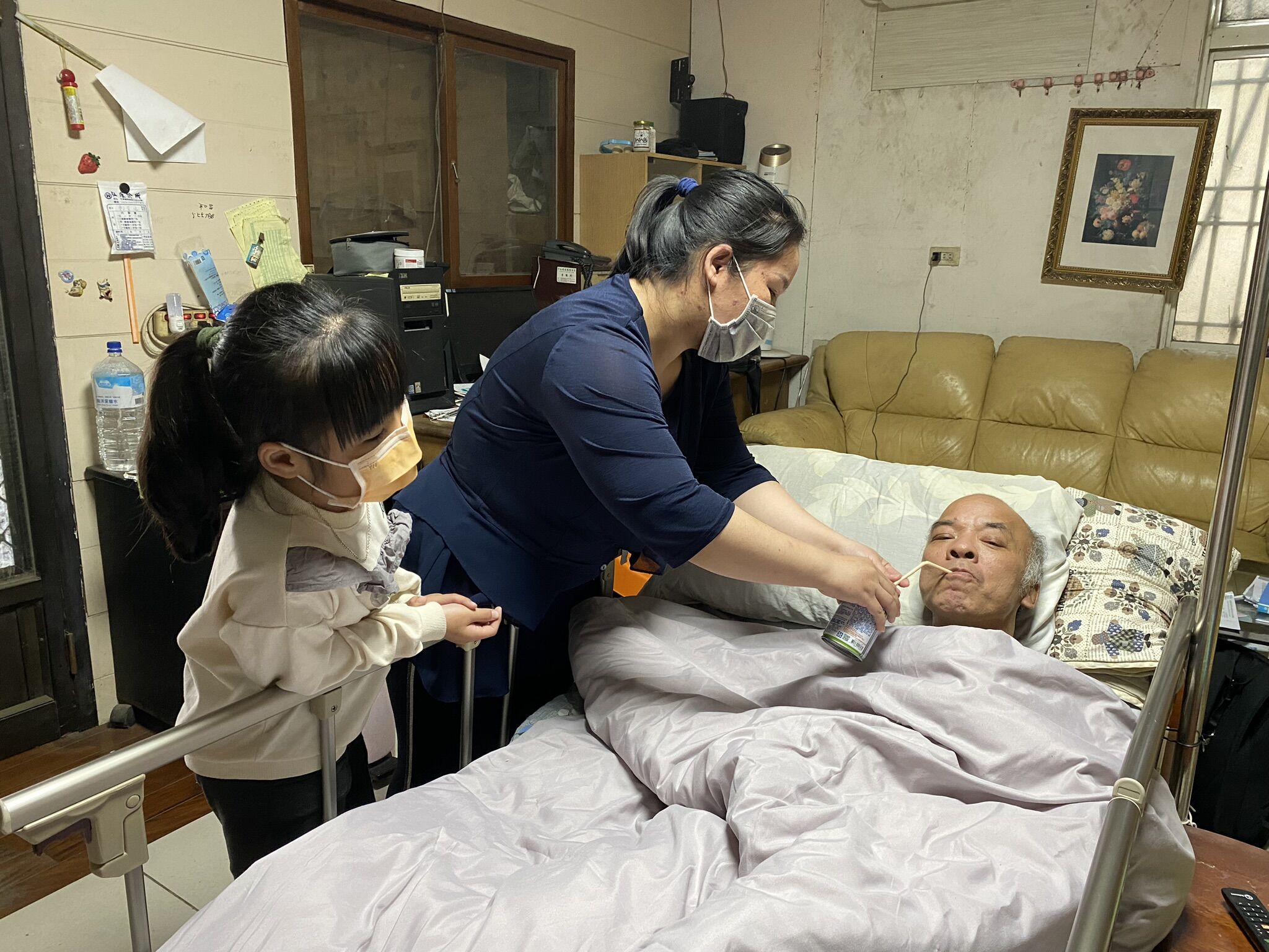 58歲阿鴻（鄭宜鴻）中風後返家，目前仍臥床，尚無法正常進食，妻子阿安（左）用吸管讓先生喝牛奶，深怕他嗆到，一旁還有7歲目前讀小2女兒小心。王雪玲攝